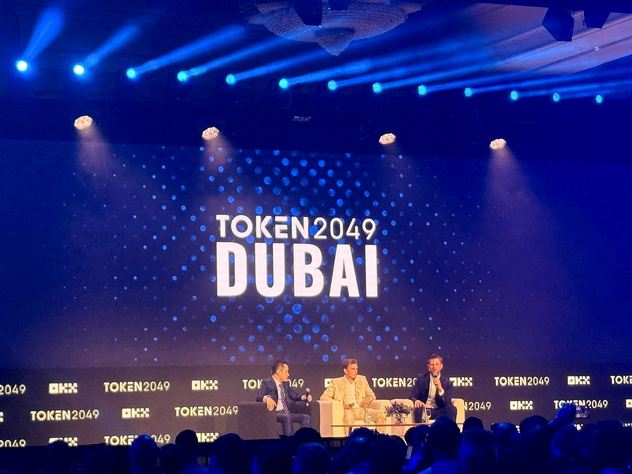 Vibrant crowd at TOKEN2049 Dubai 2026 conference hall with stablecoin USDT payment screens and travelers checking phones for bookings and onchain insurance